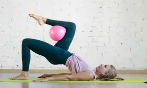 Donna esegue un esercizio di pilates con un pallina rosa tra le ginocchia, sollevando il bacino da terra.