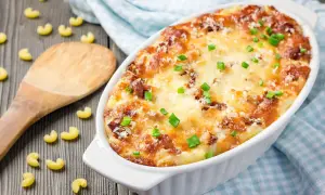 Piatto invitante di pasta al forno alla zucca, dorata e filante, guarnita con erba cipollina. Maccheroni sparsi sul tavolo di legno.