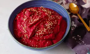 Hummus di barbabietola cremoso e vibrante, cosparso di semi di sesamo, in una ciotola blu. Un cucchiaio dorato e un tovagliolo fiorato completano la scena.