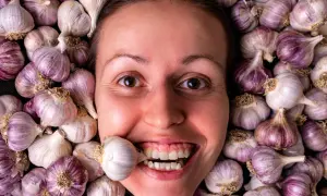 Una donna sorride con uno spicchio d'aglio tra i denti, circondata da teste d'aglio. Si dice che l'aglio fa dimagrire!