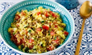 Ciotola turchese piena di cous cous con lenticchie, pomodori a cubetti e zucchine. Un cucchiaio dorato riposa su un tovagliolo blu e bianco.