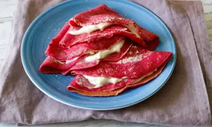 Deliziose crepes alla barbabietola, ripiene di una crema bianca e spolverate di zucchero a velo, servite su un piatto blu.