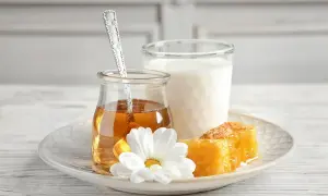 Un bicchiere di latte, un vasetto di miele dorato con un cucchiaino, e favi di miele su un piatto con un fiore bianco. Attenzione: latte e miele prima di dormire fa ingrassare.