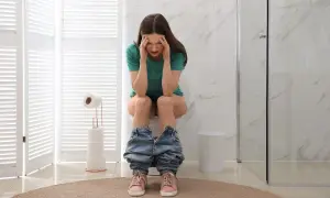 Donna con mani sulla testa, seduta sul water, con jeans abbassati. Sembra soffrire, forse ha mangiato troppo e ora feci caprine.