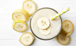 Un cremoso frullato di frutta con latte, guarnito con fette di banana e una cannuccia a righe gialle e bianche, circondato da fette di banana fresca.