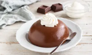 Budino con cacao amaro, guarnito con panna montata, su un piatto bianco. Accanto, due quadratini di cioccolato e una ciotola di panna.