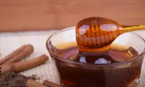 Miele di castagno ambrato, con bastoncini di cannella e polvere scura. Un cucchiaio raccoglie il miele, suggerendo benefici per l'intestino.