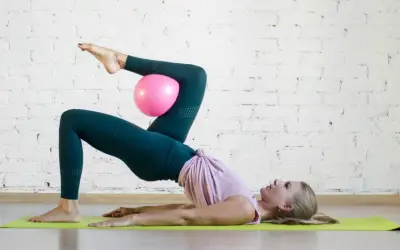 Donna esegue un esercizio di pilates con un pallina rosa tra le ginocchia, sollevando il bacino da terra.