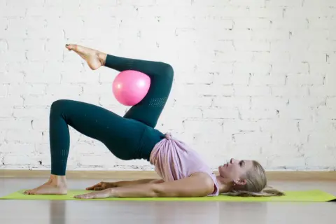 Donna esegue un esercizio di pilates con un pallina rosa tra le ginocchia, sollevando il bacino da terra.