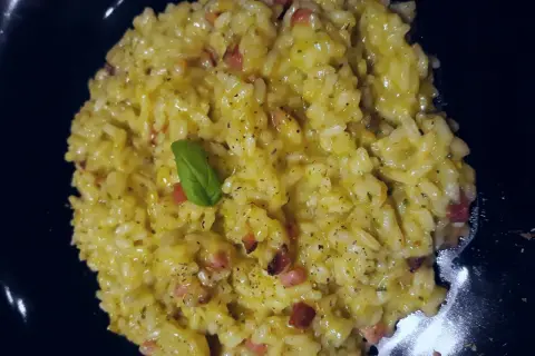 Un cremoso risotto basmati con zafferano, pancetta e una fogliolina di basilico, servito in un piatto nero.