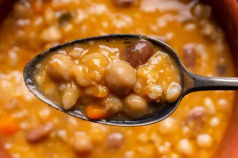 Un cucchiaio pieno di zuppa ricca di legumi e cereali, con ceci, fagioli e pezzetti di carota. Un piatto confortante e nutriente.