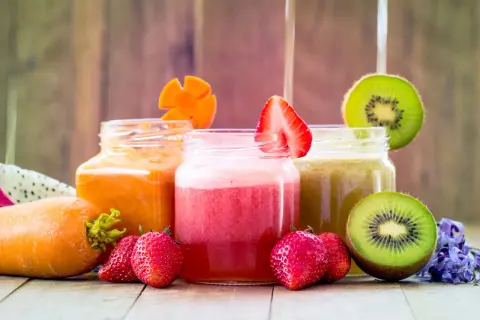 Tre vasetti di frullato di frutta colorati: arancione, rosa e verde. Fragole, kiwi e carote decorano i bicchieri.