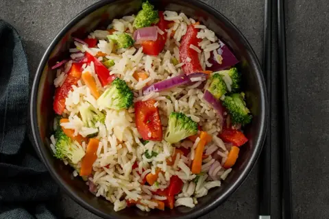 Un piatto colorato di riso con legumi e verdure miste: broccoli, peperoni rossi, carote e cipolla viola.