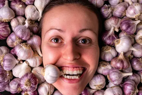 Una donna sorride con uno spicchio d'aglio tra i denti, circondata da teste d'aglio. Si dice che l'aglio fa dimagrire!