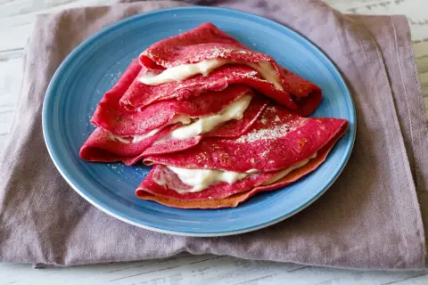 Deliziose crepes alla barbabietola, ripiene di una crema bianca e spolverate di zucchero a velo, servite su un piatto blu.