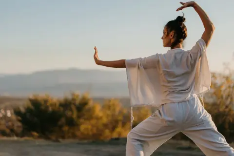 Donna pratica tai chi all'aperto, con le braccia sollevate e il corpo in posizione di equilibrio, immersa nella luce dorata del tramonto.