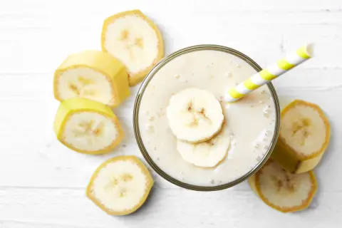 Un cremoso frullato di frutta con latte, guarnito con fette di banana e una cannuccia a righe gialle e bianche, circondato da fette di banana fresca.