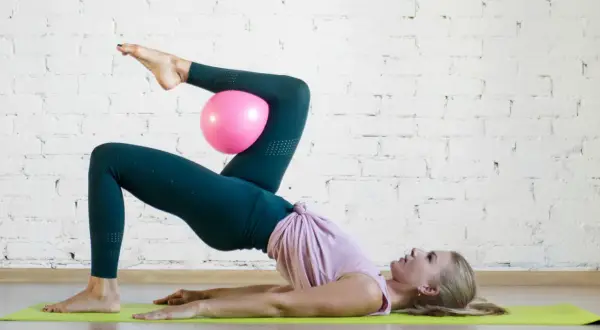 Donna esegue un esercizio di pilates con un pallina rosa tra le ginocchia, sollevando il bacino da terra.