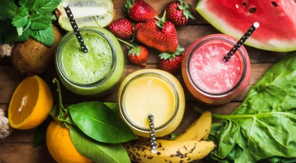 Tre colorati frullati di frutta con acqua, circondati da ingredienti freschi come fragole, kiwi, arance e spinaci.
