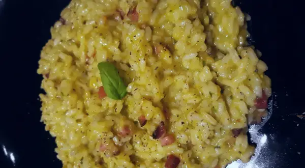 Un cremoso risotto basmati con zafferano, pancetta e una fogliolina di basilico, servito in un piatto nero.