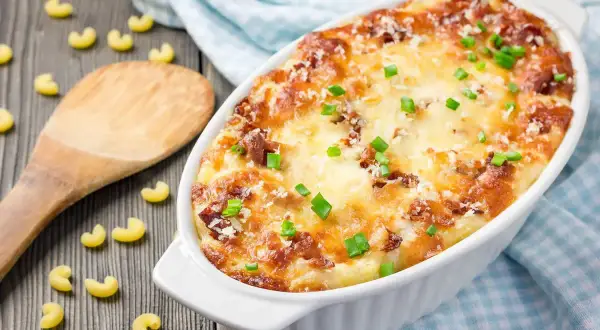 Piatto invitante di pasta al forno alla zucca, dorata e filante, guarnita con erba cipollina. Maccheroni sparsi sul tavolo di legno.