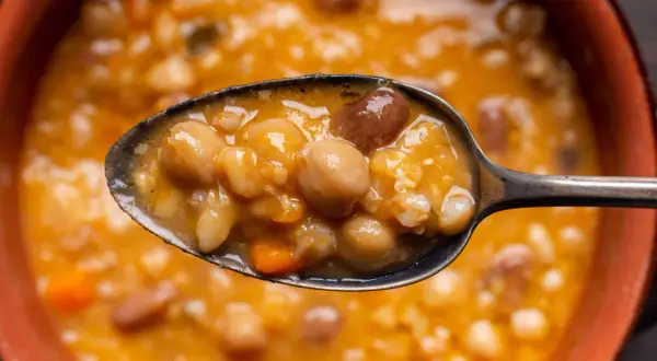 Un cucchiaio pieno di zuppa ricca di legumi e cereali, con ceci, fagioli e pezzetti di carota. Un piatto confortante e nutriente.