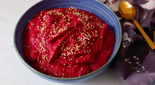 Hummus di barbabietola cremoso e vibrante, cosparso di semi di sesamo, in una ciotola blu. Un cucchiaio dorato e un tovagliolo fiorato completano la scena.