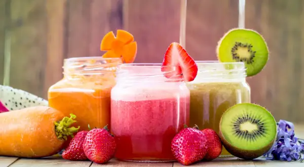 Tre vasetti di frullato di frutta colorati: arancione, rosa e verde. Fragole, kiwi e carote decorano i bicchieri.