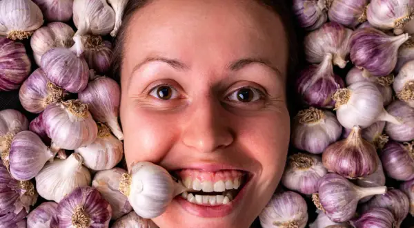Una donna sorride con uno spicchio d'aglio tra i denti, circondata da teste d'aglio. Si dice che l'aglio fa dimagrire!