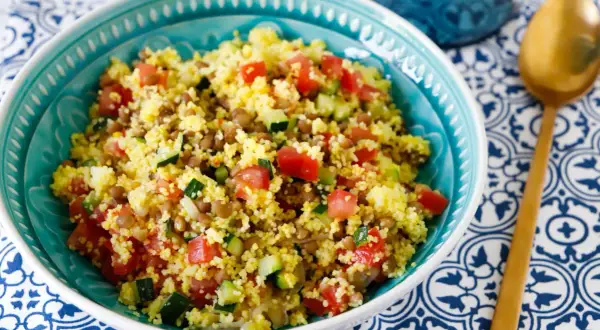 Ciotola turchese piena di cous cous con lenticchie, pomodori a cubetti e zucchine. Un cucchiaio dorato riposa su un tovagliolo blu e bianco.