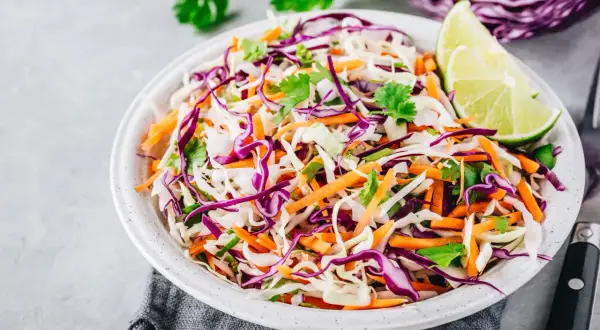 Insalate per dimagrire la pancia: cavolo cappuccio viola e bianco, carote grattugiate e coriandolo fresco, guarnite con spicchi di lime.