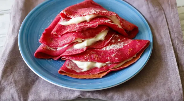 Deliziose crepes alla barbabietola, ripiene di una crema bianca e spolverate di zucchero a velo, servite su un piatto blu.