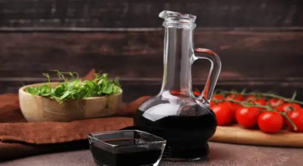 Aceto balsamico scuro in una caraffa di vetro e una ciotolina, con pomodorini e germogli sullo sfondo. Attenzione: l'aceto contiene nichel.