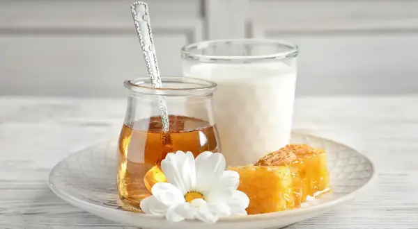 Un bicchiere di latte, un vasetto di miele dorato con un cucchiaino, e favi di miele su un piatto con un fiore bianco. Attenzione: latte e miele prima di dormire fa ingrassare.