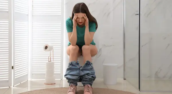Donna con mani sulla testa, seduta sul water, con jeans abbassati. Sembra soffrire, forse ha mangiato troppo e ora feci caprine.