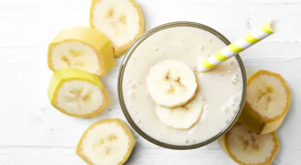 Un cremoso frullato di frutta con latte, guarnito con fette di banana e una cannuccia a righe gialle e bianche, circondato da fette di banana fresca.