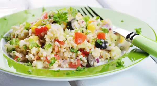 Un'insalata di miglio colorata con pomodori, olive, mais e prezzemolo fresco, servita su un piatto verde brillante.