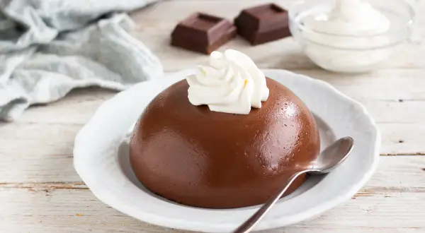 Budino con cacao amaro, guarnito con panna montata, su un piatto bianco. Accanto, due quadratini di cioccolato e una ciotola di panna.