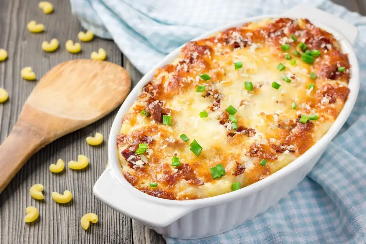 Piatto invitante di pasta al forno alla zucca, dorata e filante, guarnita con erba cipollina. Maccheroni sparsi sul tavolo di legno.