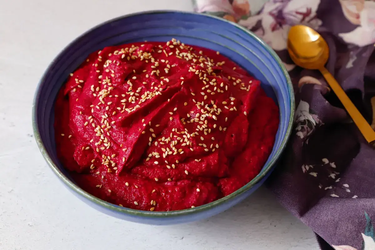 Hummus di barbabietola cremoso e vibrante, cosparso di semi di sesamo, in una ciotola blu. Un cucchiaio dorato e un tovagliolo fiorato completano la scena.