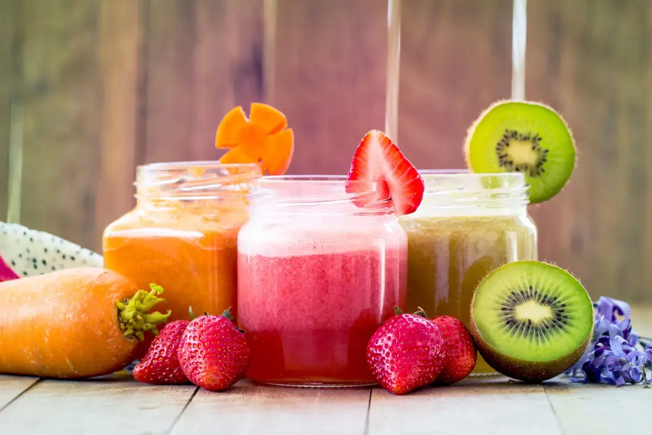 Tre vasetti di frullato di frutta colorati: arancione, rosa e verde. Fragole, kiwi e carote decorano i bicchieri.