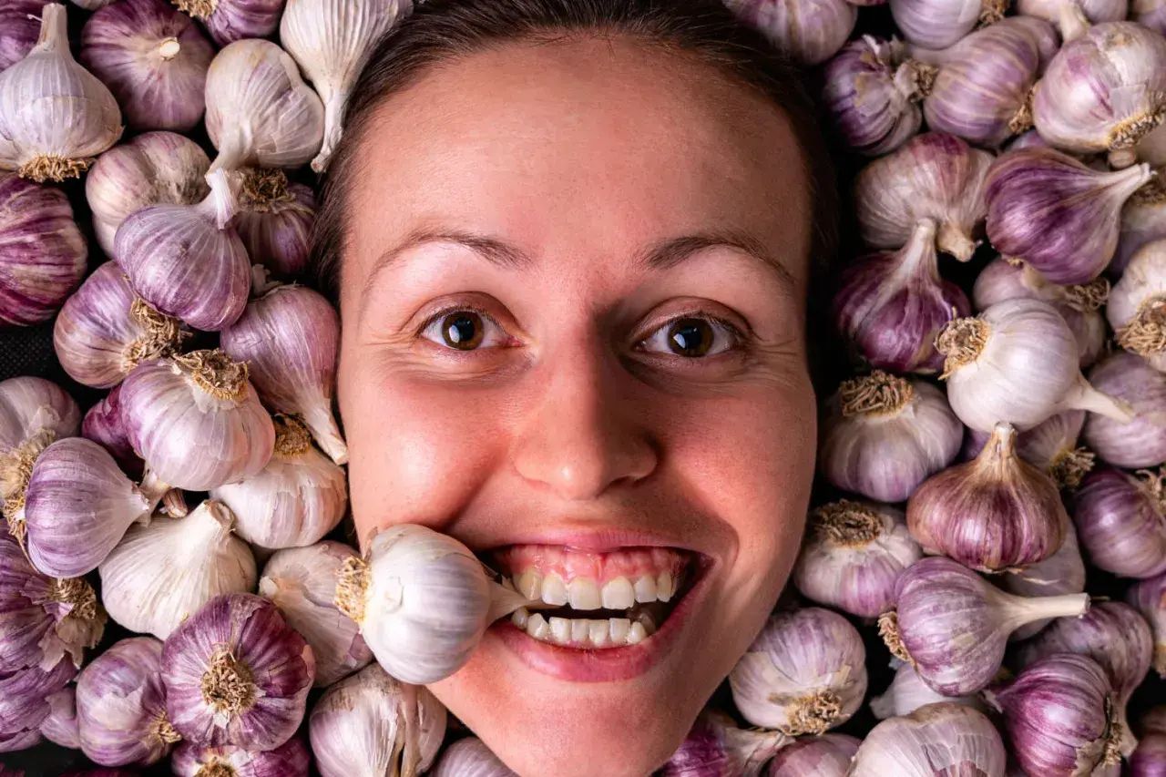 Una donna sorride con uno spicchio d'aglio tra i denti, circondata da teste d'aglio. Si dice che l'aglio fa dimagrire!
