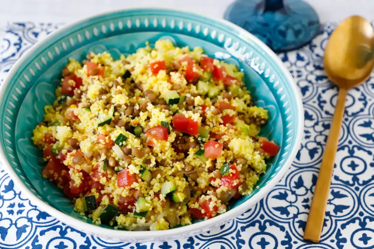 Ciotola turchese piena di cous cous con lenticchie, pomodori a cubetti e zucchine. Un cucchiaio dorato riposa su un tovagliolo blu e bianco.