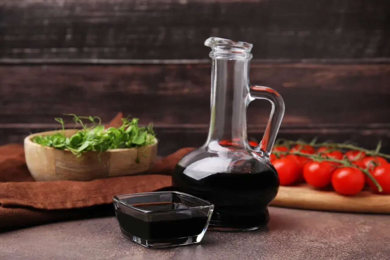 Aceto balsamico scuro in una caraffa di vetro e una ciotolina, con pomodorini e germogli sullo sfondo. Attenzione: l'aceto contiene nichel.