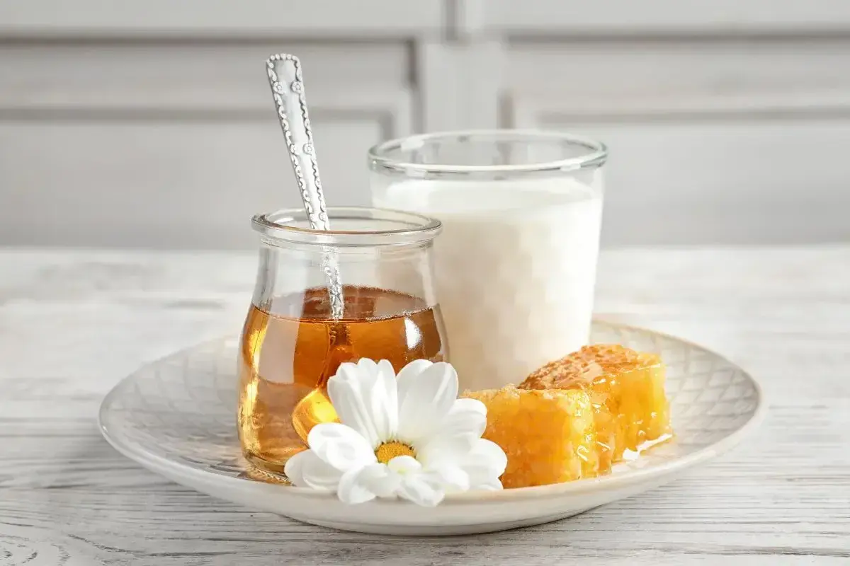 Un bicchiere di latte, un vasetto di miele dorato con un cucchiaino, e favi di miele su un piatto con un fiore bianco. Attenzione: latte e miele prima di dormire fa ingrassare.