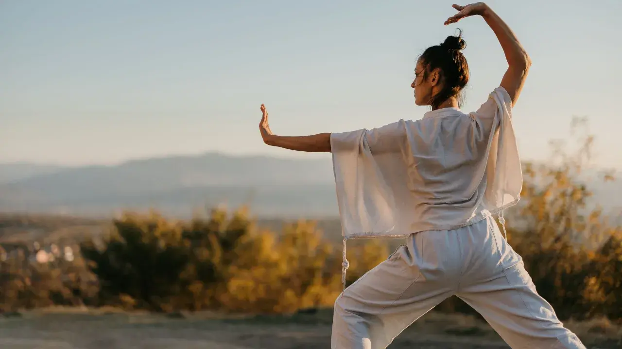 Donna pratica tai chi all'aperto, con le braccia sollevate e il corpo in posizione di equilibrio, immersa nella luce dorata del tramonto.