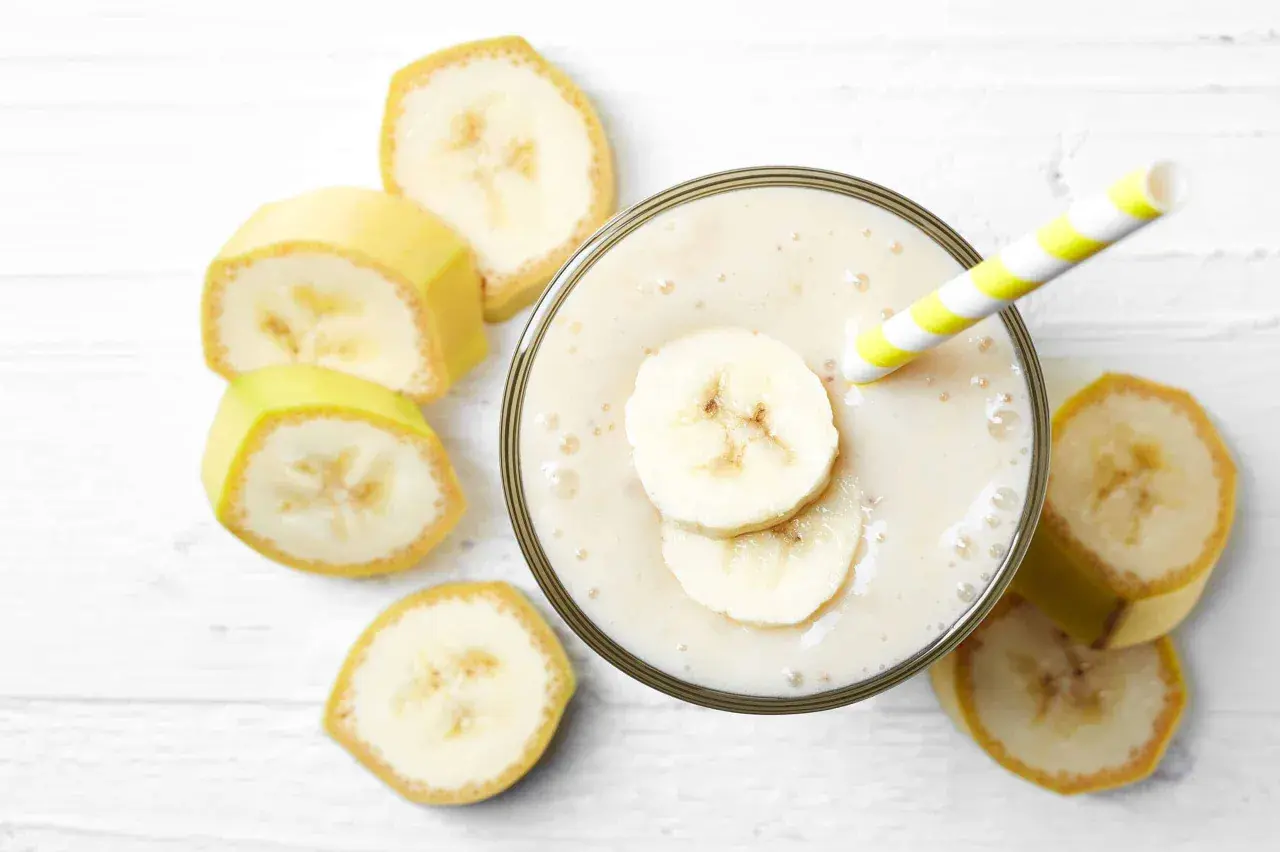 Un cremoso frullato di frutta con latte, guarnito con fette di banana e una cannuccia a righe gialle e bianche, circondato da fette di banana fresca.
