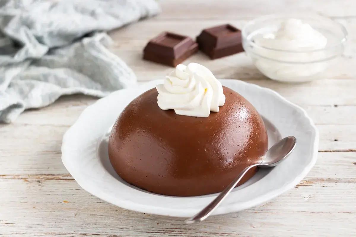 Budino con cacao amaro, guarnito con panna montata, su un piatto bianco. Accanto, due quadratini di cioccolato e una ciotola di panna.
