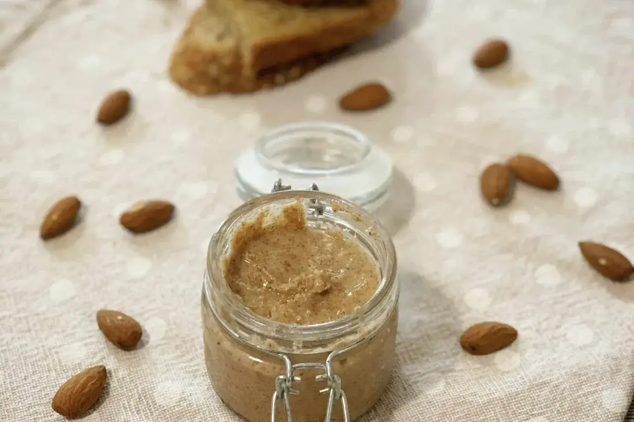Barattolo di crema di mandorle, perfetta per tante ricette. Mandorle sparse intorno, con una fetta di pane in lontananza.