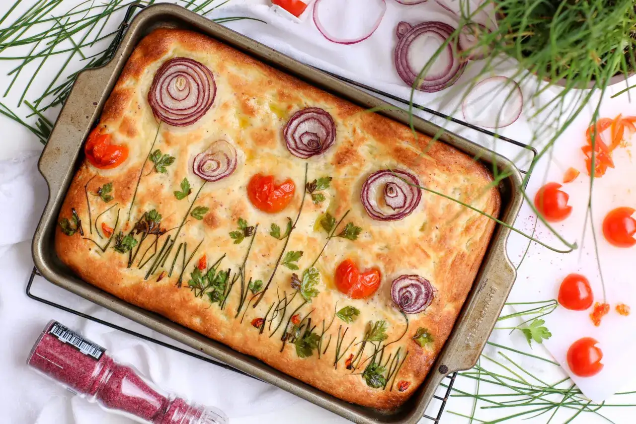 Focaccia di verdure decorata con cipolle rosse, pomodorini e erbe aromatiche, che ricorda un prato fiorito.